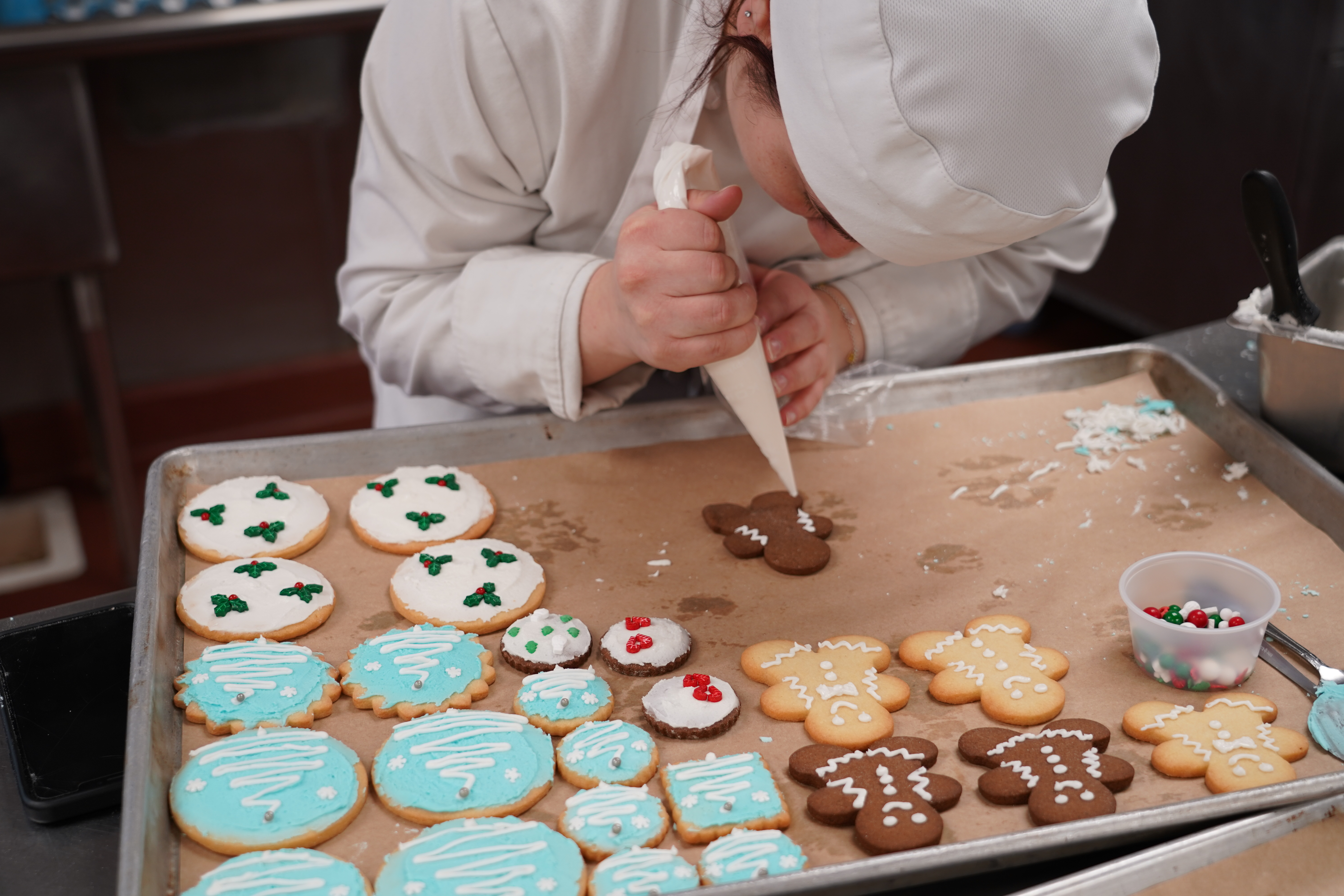 A student pipes royal icing onto a cookie on a sheet pan of decorated cookies