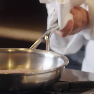 A chef in a white coat cooks on a stove with a stainless steel pan with sloped sides