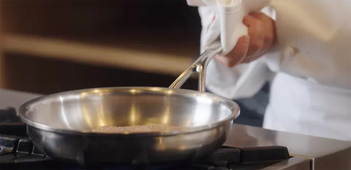 A chef in a white coat cooks on a stove with a stainless steel pan with sloped sides
