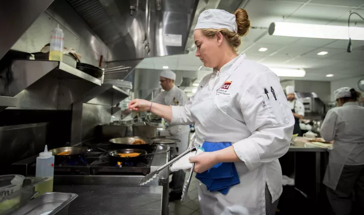 A culinary arts student seasons food in a saute pan on a stove at culinary school